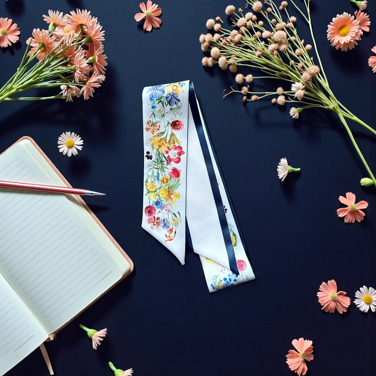 White floral handbag twilly scarf featuring colourful garden blooms and navy edging, arranged beside a notebook and pink flowers.