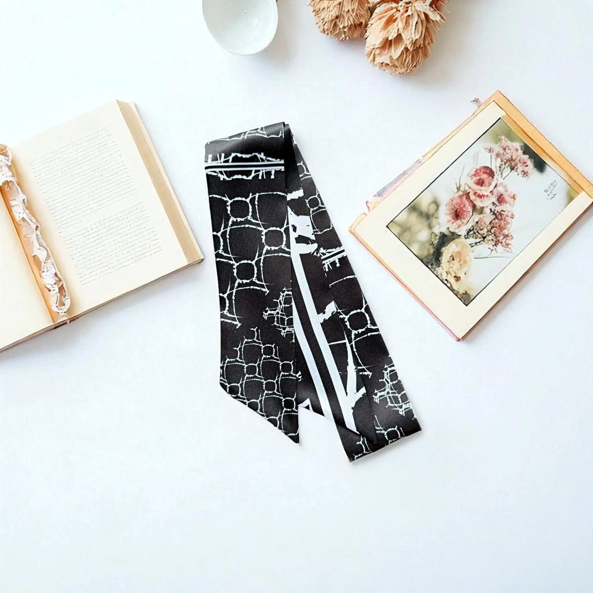Black geometric skinny scarf with contrasting white striped details, arranged beside an open book and framed botanical artwork.