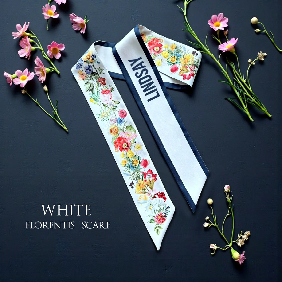 Personalised white twilly scarf featuring colourful watercolour wildflowers, navy blue trim, and custom name Lindsay, displayed on a dark background.