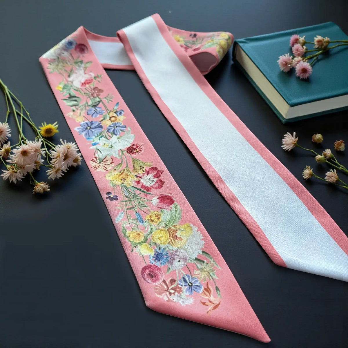 Romantic pink skinny scarf featuring delicate roses, daisies, and colourful garden blooms, displayed beside books and dried flowers.