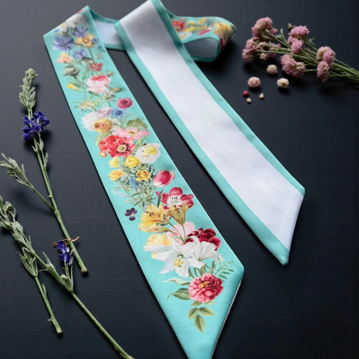 Elegant turquoise skinny scarf with colourful floral illustrations and a white reverse side, displayed with dried flowers.