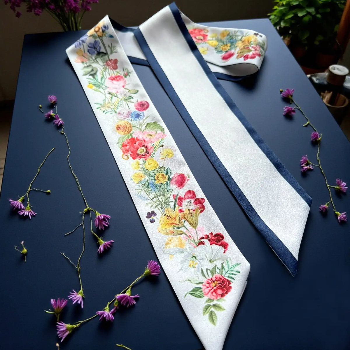 Elegant white skinny scarf with vibrant botanical flowers, a classic navy border, and personalised lettering, styled with delicate flowers.