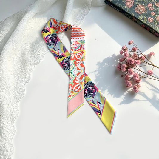 A colorful patterned twilly scarf laid flat on a white surface, styled with a white lace cloth, dried pink flowers, and a floral book.
