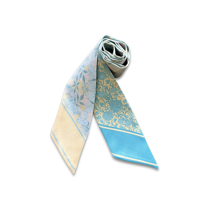 Pastel blue floral twilly scarf with vintage ornamental pattern shown partially rolled on a neutral background