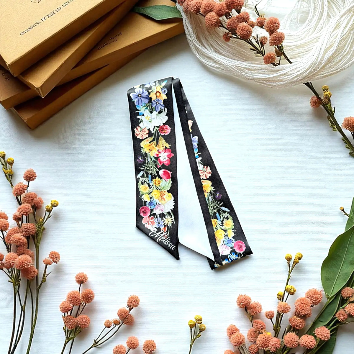 Personalised black botanical skinny scarf decorated with colourful watercolour flowers, styled on a white surface with dried floral arrangements