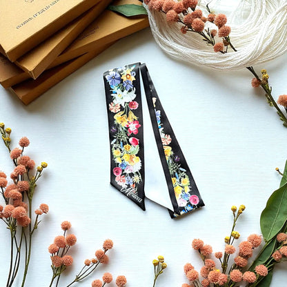 Personalised black botanical skinny scarf decorated with colourful watercolour flowers, styled on a white surface with dried floral arrangements