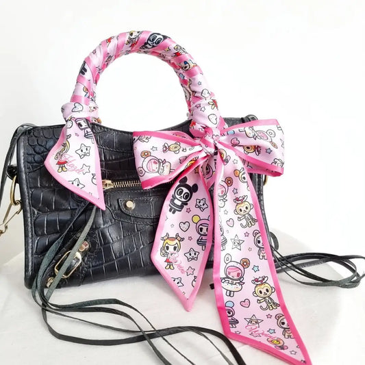 Pink satin twilly scarf featuring cute kawaii cartoon characters, stars, and hearts, wrapped around the handle of a black crocodile-textured handbag with a decorative bow.