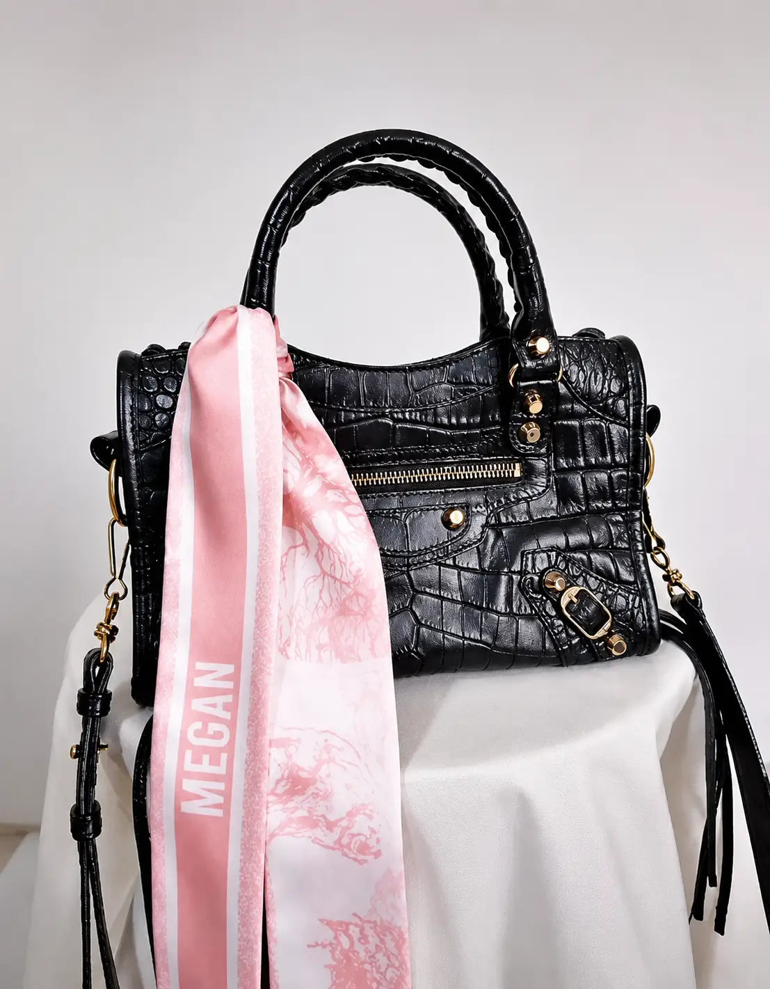 Personalised pink satin twilly scarf featuring the name Megan in white lettering, tied around the handle of a black crocodile-textured handbag.