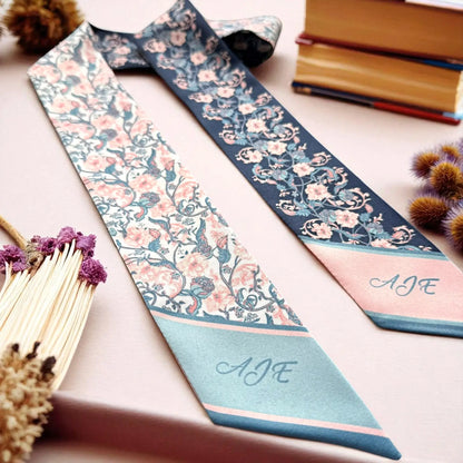Ivory satin twilly scarf featuring a vintage-inspired botanical pattern with pink flowers and intricate blue-green vines, accented by a turquoise pointed end with custom initials.
