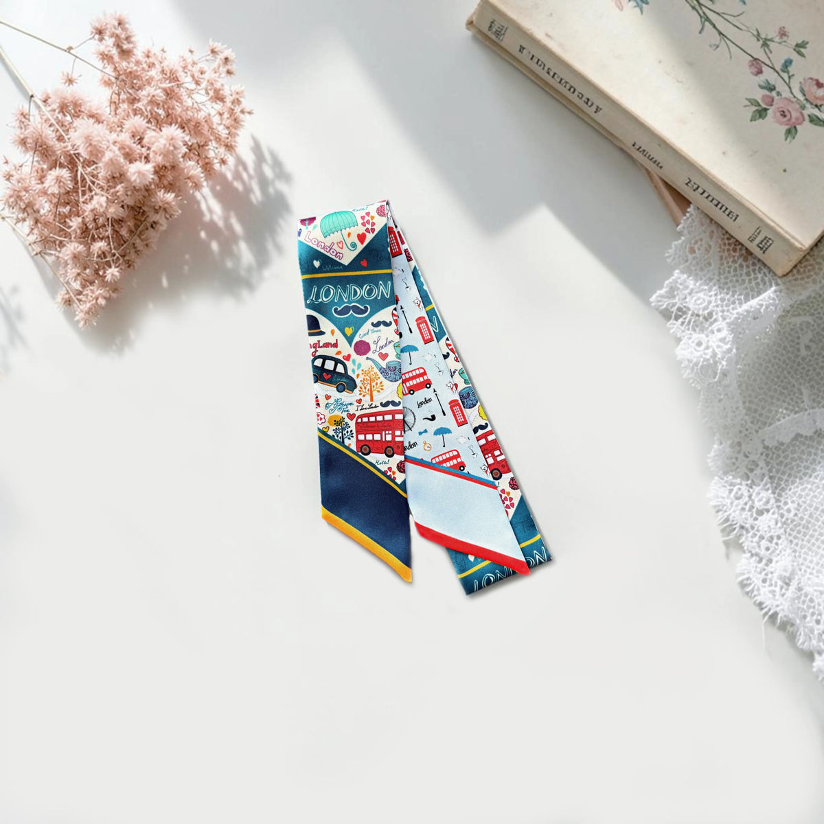 Playful London novelty skinny scarf with red double-decker buses, black taxis, famous landmarks, and colorful illustrations, suitable for styling as a hair scarf or handbag accessory.