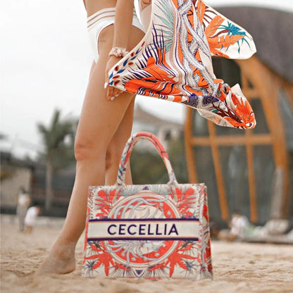 Structured personalized canvas book tote with tropical botanical artwork and embroidered name, carried on a beach.