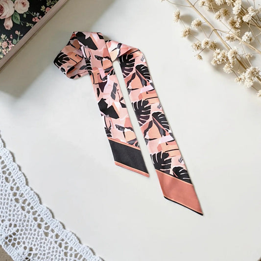 twilly scarf featuring bold black tropical monstera leaves, abstract botanical patterns, and contrasting borders, elegantly arranged on a soft neutral background.