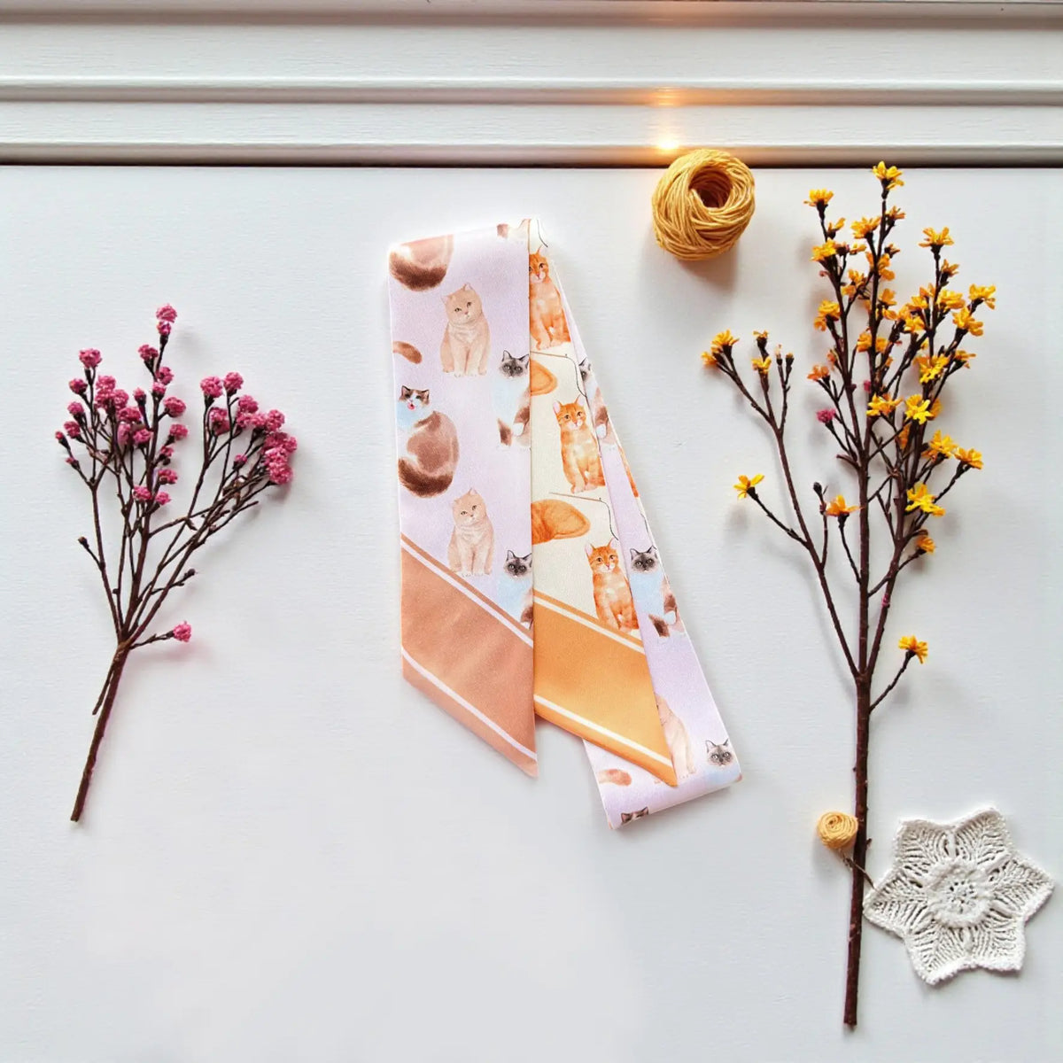 Whimsical cat twilly scarf featuring repeating feline illustrations in pastel pink, cream and orange shades.