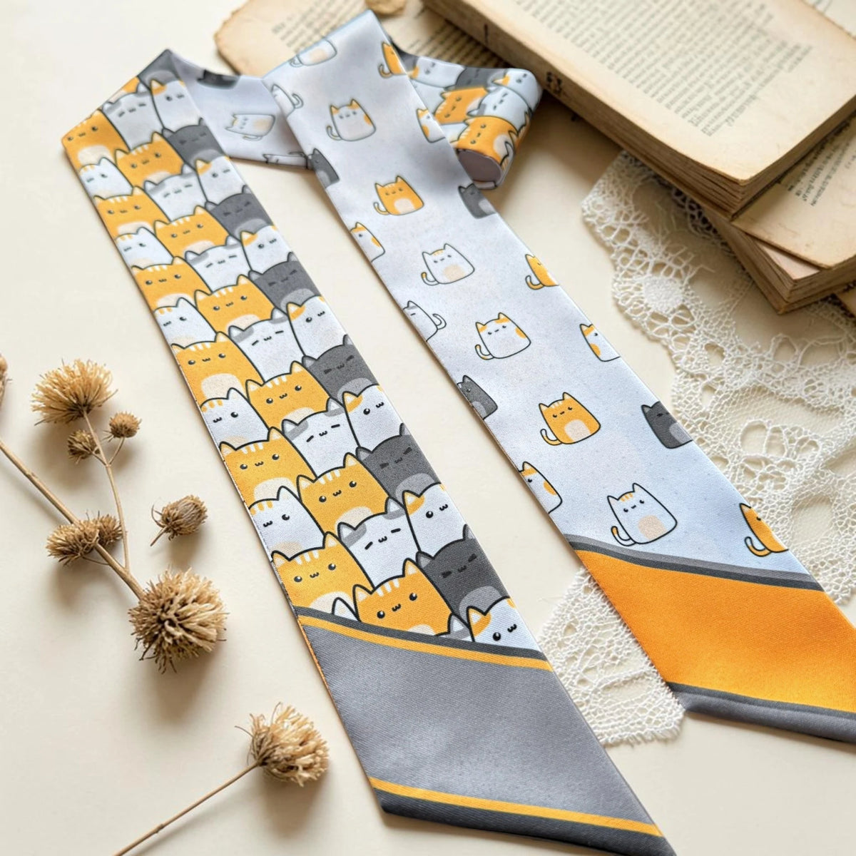 Whimsical cat pattern twilly scarf displayed with books and lace, featuring playful orange, white and grey cats.