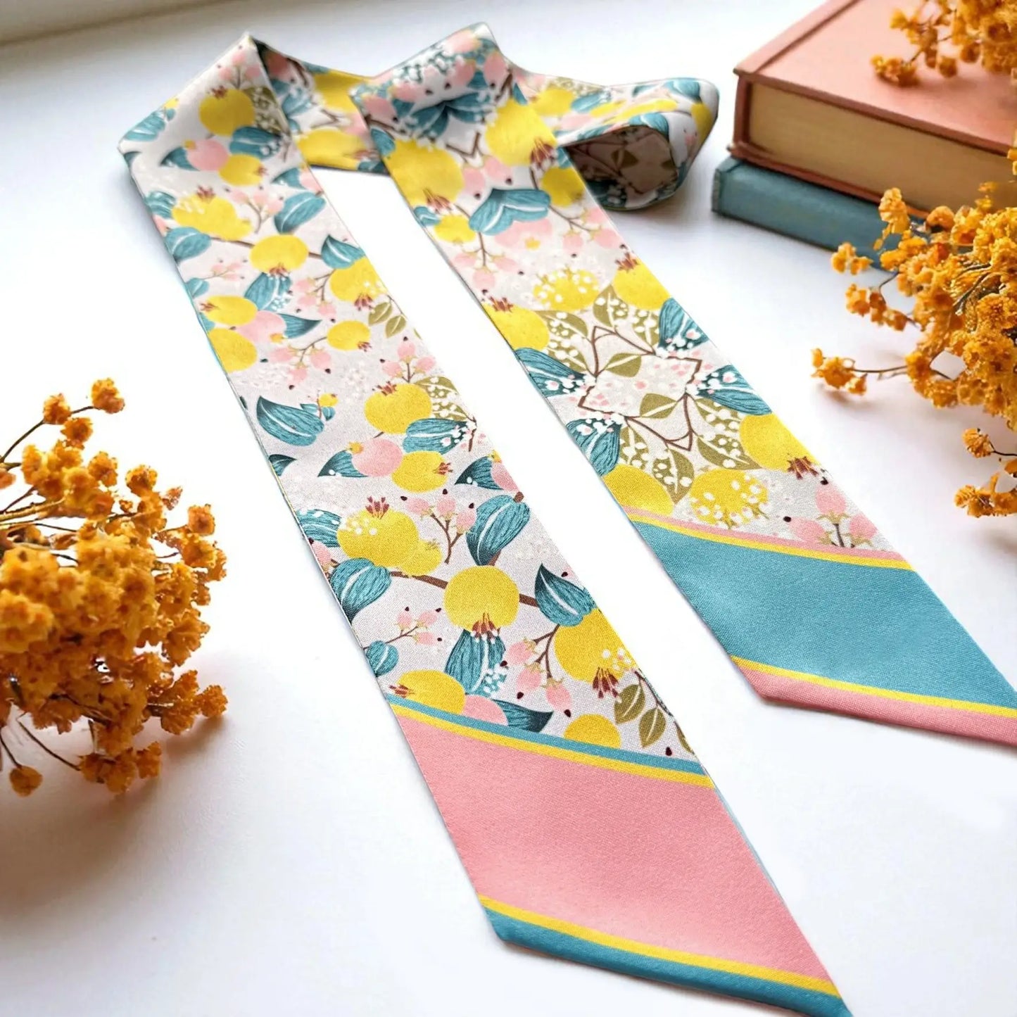 A floral silk necktie with yellow lemons, green leaves, and pink and teal stripes, styled on a white surface with dried yellow flowers and books.