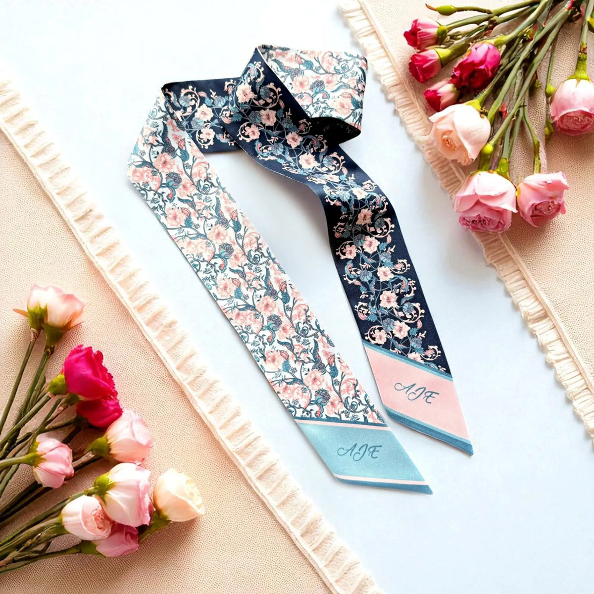 Personalised double-sided satin twilly scarf featuring delicate pink flowers, intricate blue botanical vines, and custom initials AJE on pastel-coloured pointed ends.