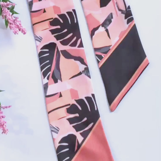 Close-up video showcasing a peach pink and black tropical leaf twilly scarf, highlighting its bold monstera pattern, smooth satin-like fabric, contrasting pointed ends, and elegant handmade finish.