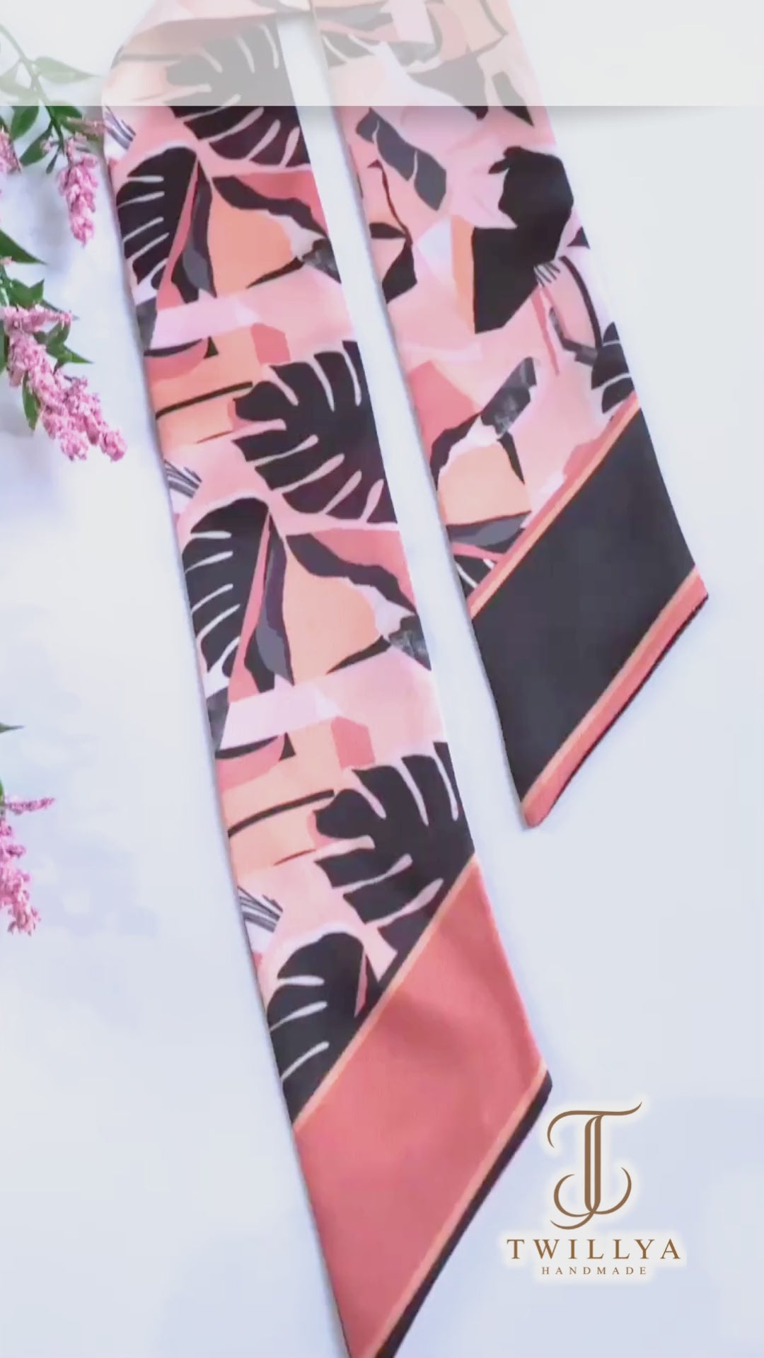 Close-up video showcasing a peach pink and black tropical leaf twilly scarf, highlighting its bold monstera pattern, smooth satin-like fabric, contrasting pointed ends, and elegant handmade finish.