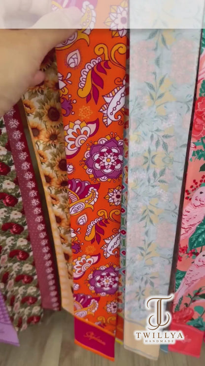 Colorful bohemian twilly scarf showcasing intricate mandala-inspired flowers, orange paisley patterns, and vibrant turquoise geometric details. Finished with personalized name printing on contrasting pink and coral pointed ends, this versatile skinny scarf adds a playful retro touch to handbags, hairstyles, and everyday outfits.