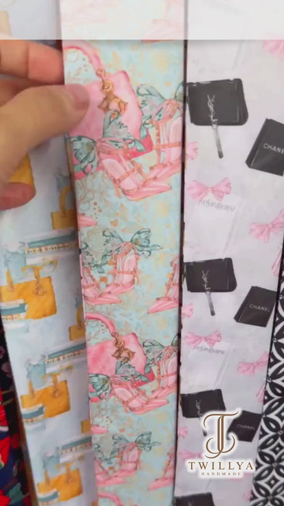 Personalized satin twilly scarves featuring pastel pink and turquoise designs with beauty-inspired motifs, including lipstick, makeup brushes, perfume bottles, and handbags. Each scarf is finished with the custom name "Tracey" on its pointed end and styled elegantly on a soft cream background.