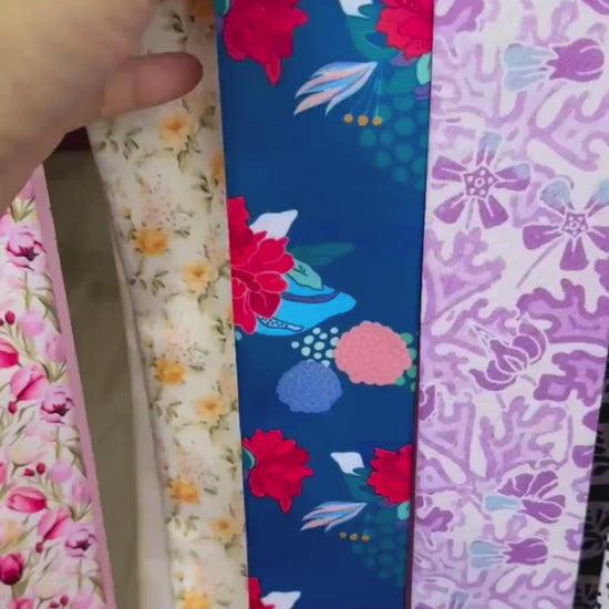 A personalised double-sided satin twilly scarf showcasing two contrasting patterns: colourful floral illustrations on a deep blue background and an intricate tan geometric print. Both pointed ends feature the custom name Elisabeth. The scarf can be tied around handbag handles, styled as a hair accessory, or worn around the neck.